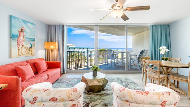 A spacious living room with a red sofa, patterned armchairs, a wooden coffee table, and a ceiling fan. A large window offers a view of the balcony and the ocean. - Sandcastle Condominiums Unit 212 (Port Aransas)