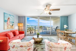 A spacious living room with a red sofa, patterned armchairs, a wooden coffee table, and a ceiling fan. A large window offers a view of the balcony and the ocean. - Sandcastle Condominiums Unit 212 (Port Aransas)