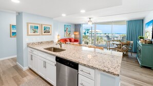 A modern kitchen with a granite countertop, stainless steel dishwasher, and white cabinetry. There is a dining area with a table and chairs, and a balcony with a view of the ocean. - Sandcastle Condominiums Unit 212 (Port Aransas)