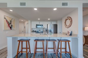 Una cocina moderna con armarios blancos, un refrigerador de acero inoxidable y una encimera de granito. Hay tres taburetes de bar de madera y un adorno decorativo en la pared.