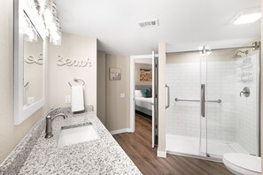 A bathroom with a granite countertop, a white sink, a towel rack, a glass shower enclosure, and a door leading to a bedroom. - Sandcastle Condominiums Unit 313 (Port Aransas)