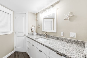 A bathroom with a granite countertop, white cabinets, a large mirror, and starfish decorations. - Sandcastle Condominiums Unit 315 (Port Aransas)