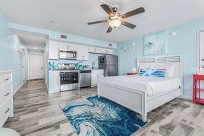 A bedroom with a bed, a ceiling fan, and a kitchen area with cabinets and appliances.