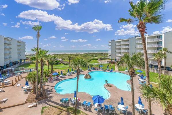 A resort pool area with lounge chairs and umbrellas, surrounded by palm trees and a view of the ocean.
