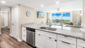 A modern kitchen with white cabinets, granite countertops, and a dishwasher. There is a large window with a view of the outdoors. - Sandcastle Condominiums Unit 323 (Port Aransas)