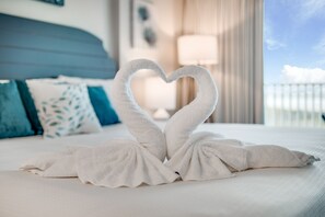 A bed with a heart-shaped towel arrangement, a blue headboard, and a view of the sky through a window. - Sandcastle Condominiums Unit 406 (Port Aransas)