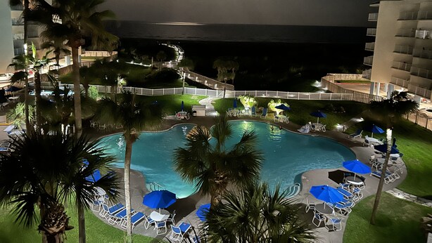 Vue nocturne d’un espace de piscine d’un complexe hôtelier, avec une piscine, des chaises longues, des parasols et des palmiers.