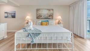 A bedroom with a white bed, two bedside tables with lamps, a painting above the bed, and a view of the outdoors through large windows. - Sandcastle Condominiums Unit 527 (Port Aransas)