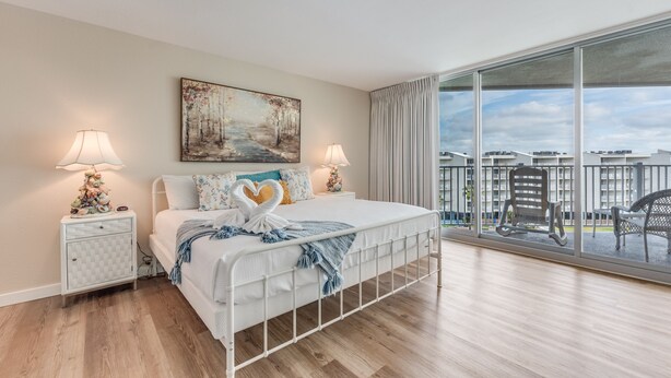 A modern bedroom with a large bed, two bedside tables with lamps, a painting on the wall, and a balcony with outdoor furniture. - Sandcastle Condominiums Unit 527 (Port Aransas)