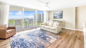 Ein geräumiges Wohnzimmer mit einem großen Schiebeglasfenster, das auf einen Balkon führt, einem beige farbenen Sofa, einem gemusterten Teppich und einem Deckenventilator.