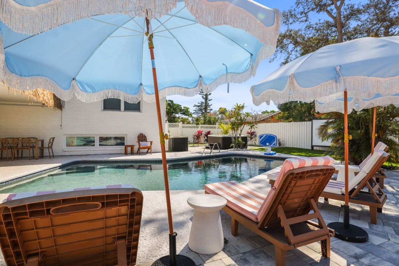 Une piscine entourée de chaises longues et de parasols, elle-même bordée d’une maison blanche et d’une cour clôturée.