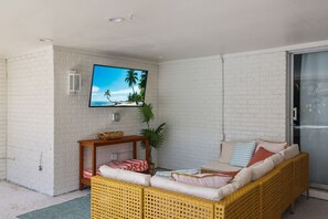 Une pièce avec un mur en briques blanches, un téléviseur fixé au-dessus d’une console en bois, un canapé en osier avec des coussins et une plante en pot.