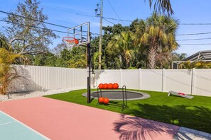 A basketball court with a hoop, a trampoline, and a white fence.
