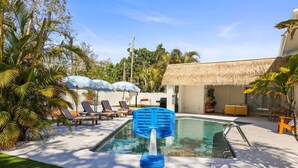 A swimming pool with lounge chairs and umbrellas, surrounded by palm trees and a thatched-roof building.
