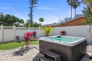 Un bain à remous dans un jardin, avec une terrasse en bois, une clôture blanche et diverses plantes.