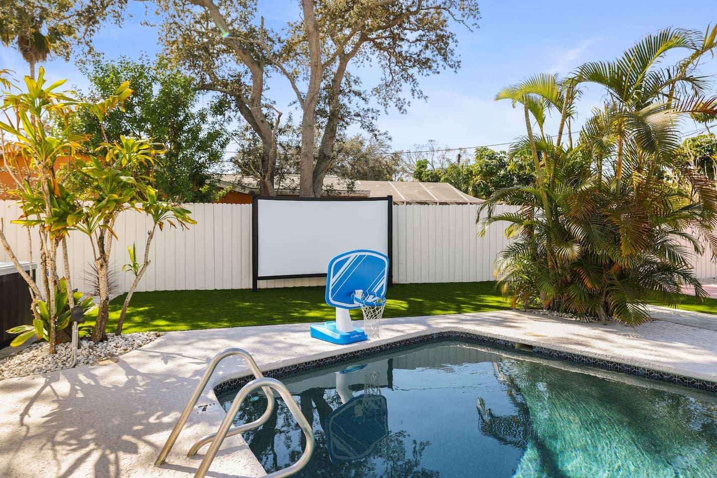 Ein Swimmingpool mit Sprungbrett in Blau und Weiß, umgeben von verschiedenen tropischen Pflanzen und einem weißen Zaun.