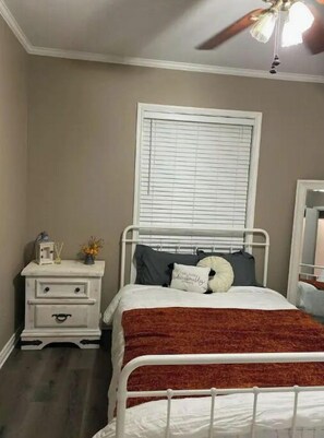 Cozy retreat with a comfortable white bed, nightstand, and dresser, plus a window with blinds and a ceiling fan.