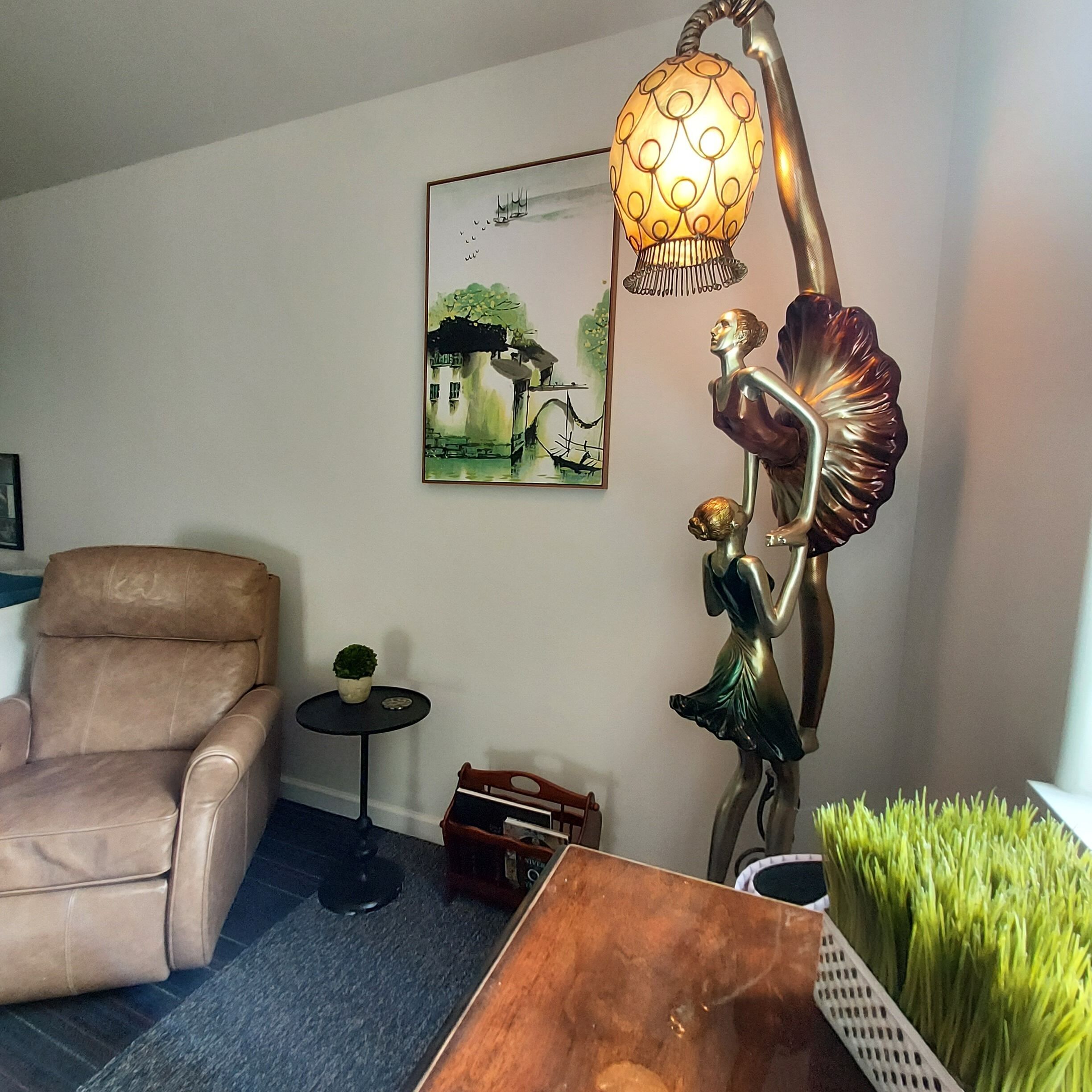 A living room with a brown leather armchair, a small round table with a potted plant, a wooden coffee table, and a wall-mounted lamp with a decorative figure.