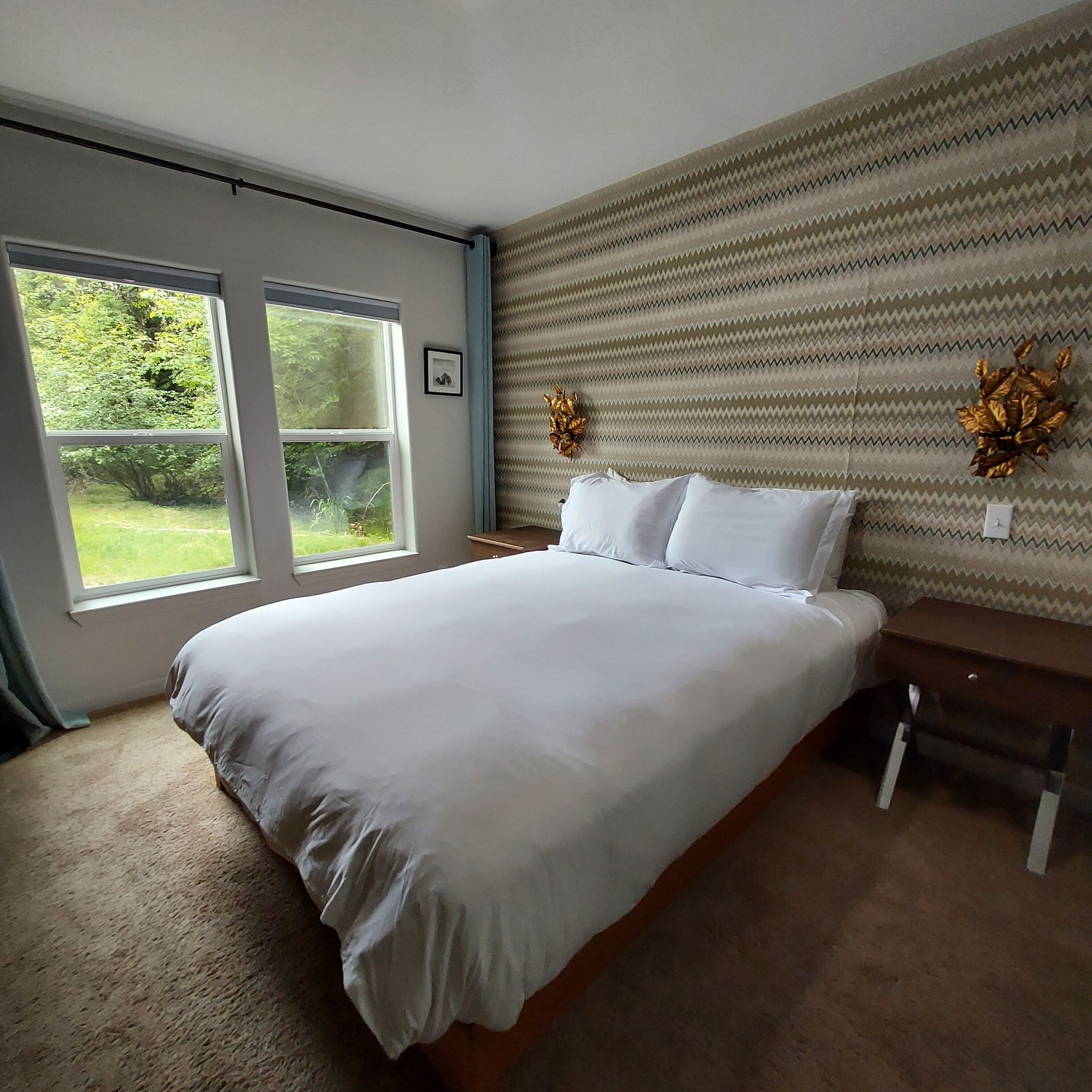 Quarto com cama grande, duas janelas, cabeceira estampada e uma pequena mesa de cabeceira.
