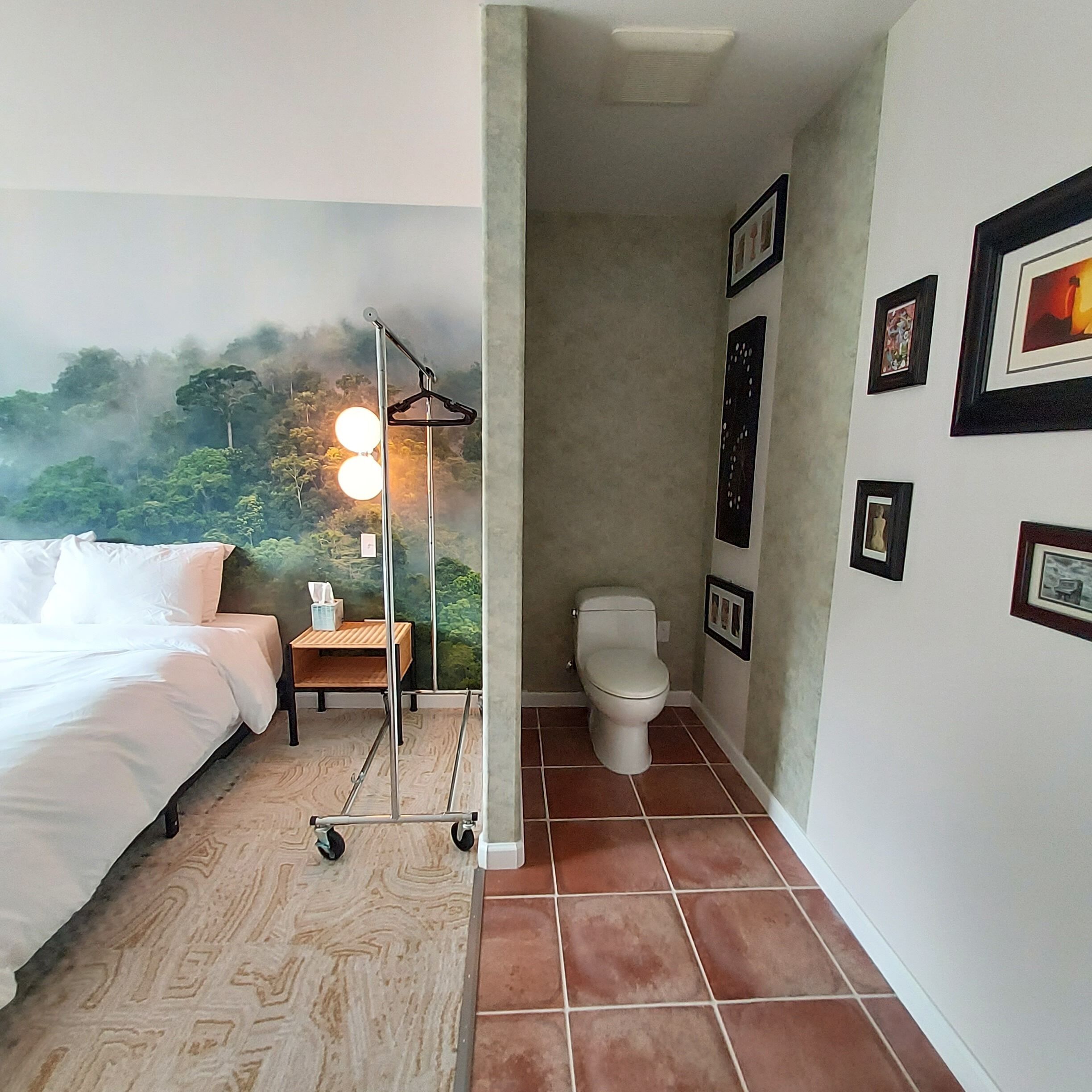 Une chambre d’hôtel comprenant un lit, une table de chevet, des toilettes fixées au mur et une salle de bain carrelée.