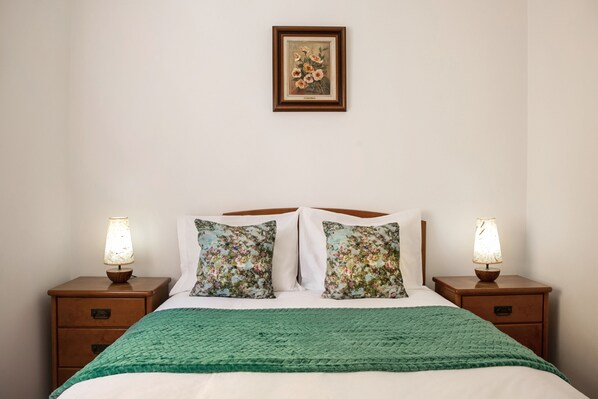 A neatly made bed with a green blanket, two floral pillows, and two bedside lamps on wooden nightstands.
