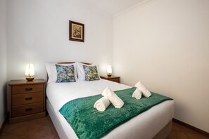A neatly made bed with a green blanket, two pillows, and two rolled towels. There are two bedside tables with lamps and a framed picture on the wall.