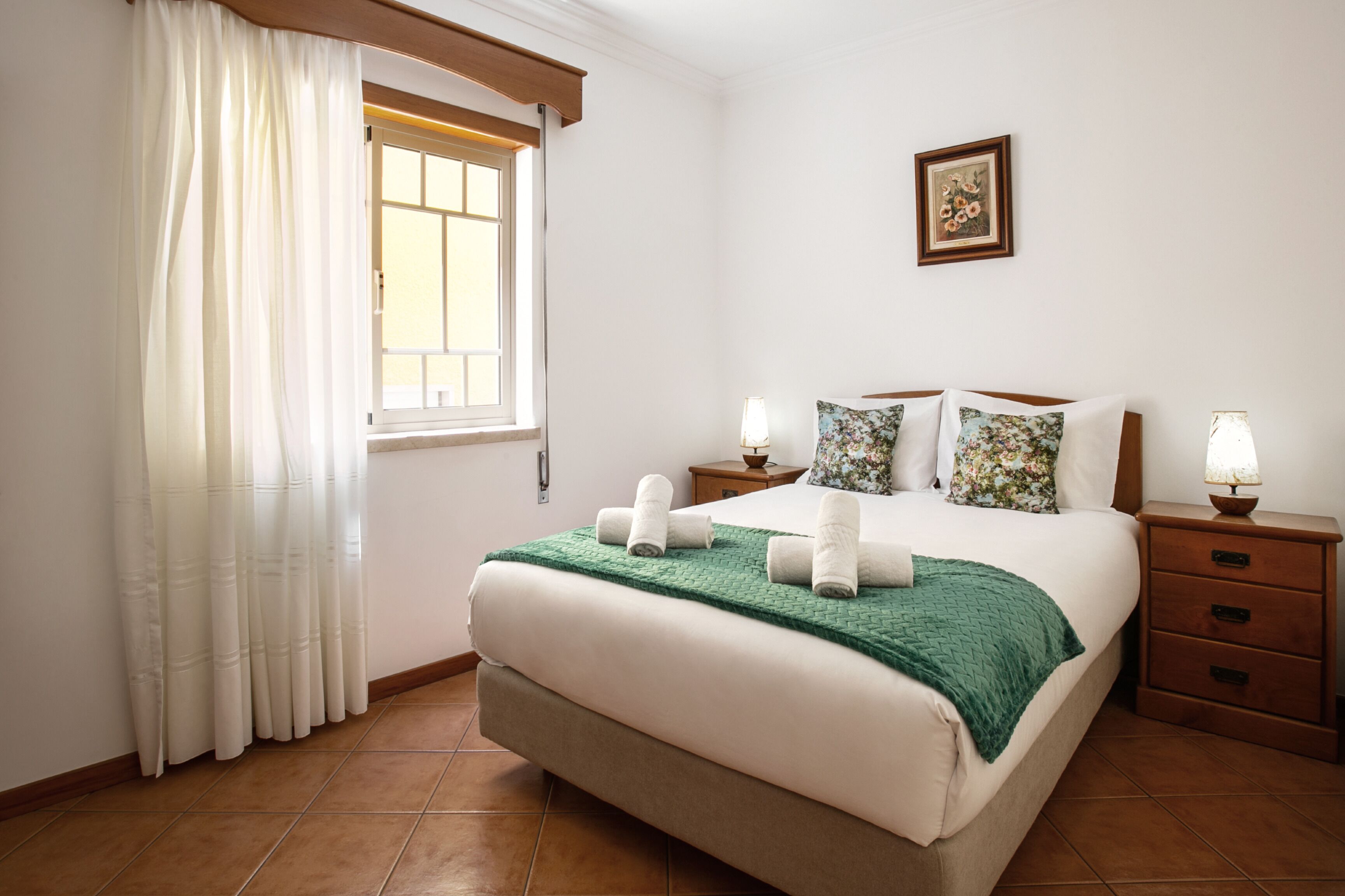 A neatly made bed with a green blanket and white pillows, a bedside table with a lamp, a window with white curtains, and a framed picture on the wall.