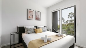 Habitación de hotel moderna con una cama grande, una mesita de noche, una silla y un ventanal que ofrece vistas a árboles.