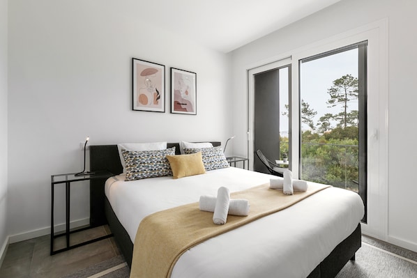 A modern hotel room with a large bed, a bedside table, a chair, and a large window offering a view of trees.