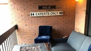 Un balcón con un muro de ladrillo, un cartel que dice 'APRES SKI' y otro que indica 'SOLO EXPERTOS'. Hay una mesa de metal negro con un brasero, un sofá gris y sillas azules.