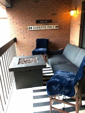 Un balcón con un muro de ladrillo, un cartel que dice 'APRES SKI' y otro que indica 'SOLO EXPERTOS'. Hay una mesa de metal negro con un brasero, un sofá gris y sillas azules.