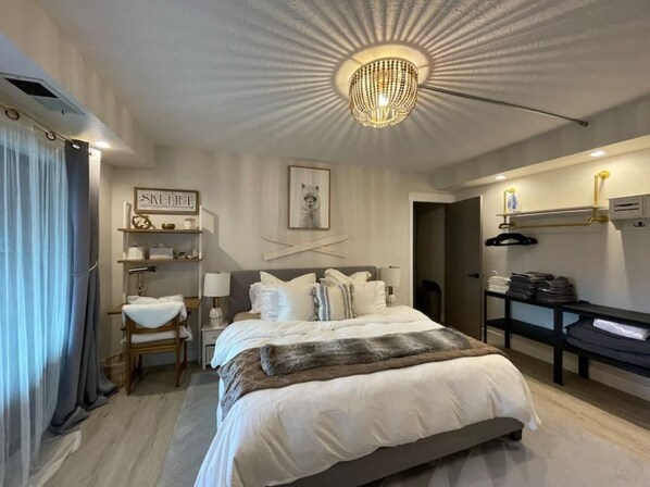 A bedroom with a large bed, a chandelier, a bedside table, a chair, a wardrobe, and a wall-mounted shelf.