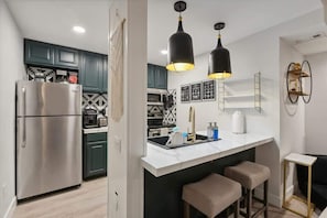 A modern kitchen with a stainless steel refrigerator, dark green cabinets, a white countertop, two bar stools, and pendant lighting.