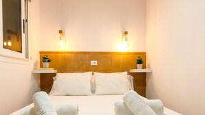 Una cama bien hecha con sábanas blancas y dos almohadas, flanqueada por dos mesitas de noche con plantas en macetas, debajo de un cabecero de madera con luces fijadas a la pared.