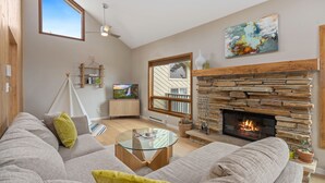 A modern living room with a stone fireplace, a glass coffee table, and a large window providing natural light. - Cliffview Luxury Vacation Rental at Windcliff (Estes Park)