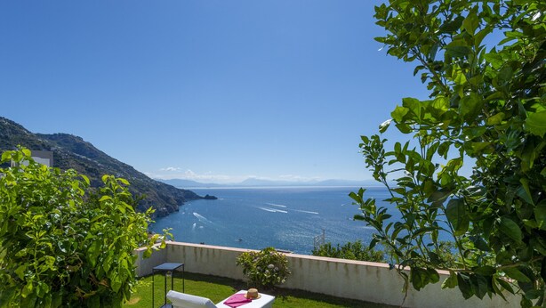 Eine Terrasse mit zwei weißen Liegestühlen, einem kleinen Tisch mit Getränken und Blick auf das Meer und die Berge.