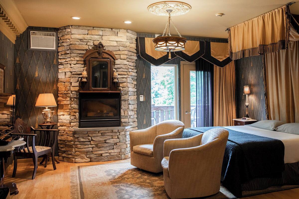 A cozy bedroom with a stone fireplace, a bed, a chandelier, and a view of the outdoors.
