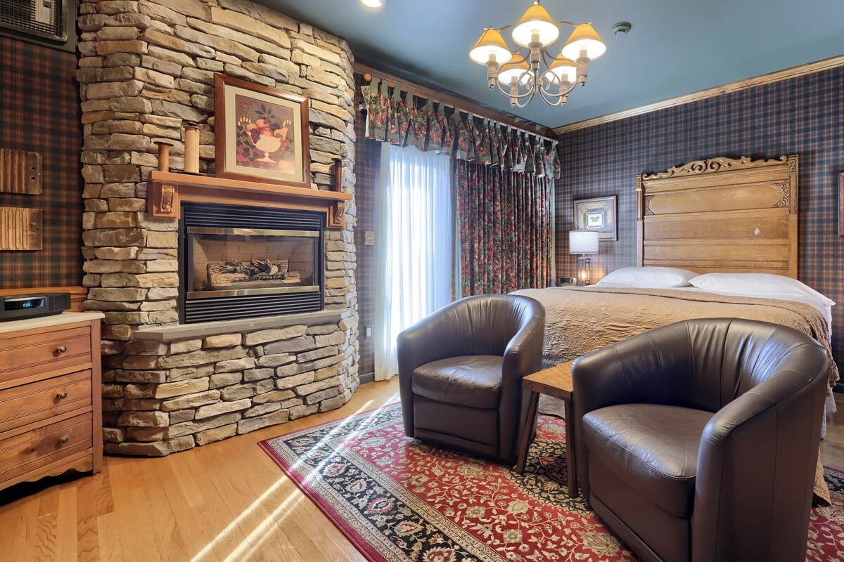 A bedroom with a stone fireplace, a large bed, leather armchairs, and a chandelier.