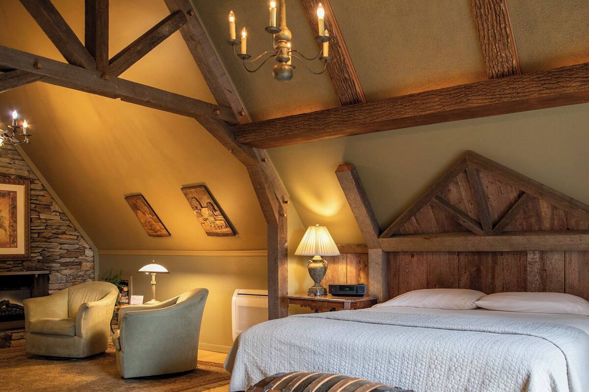 A cozy attic bedroom with exposed wooden beams, a stone fireplace, a bed with a white quilt, and a stone wall with artwork.