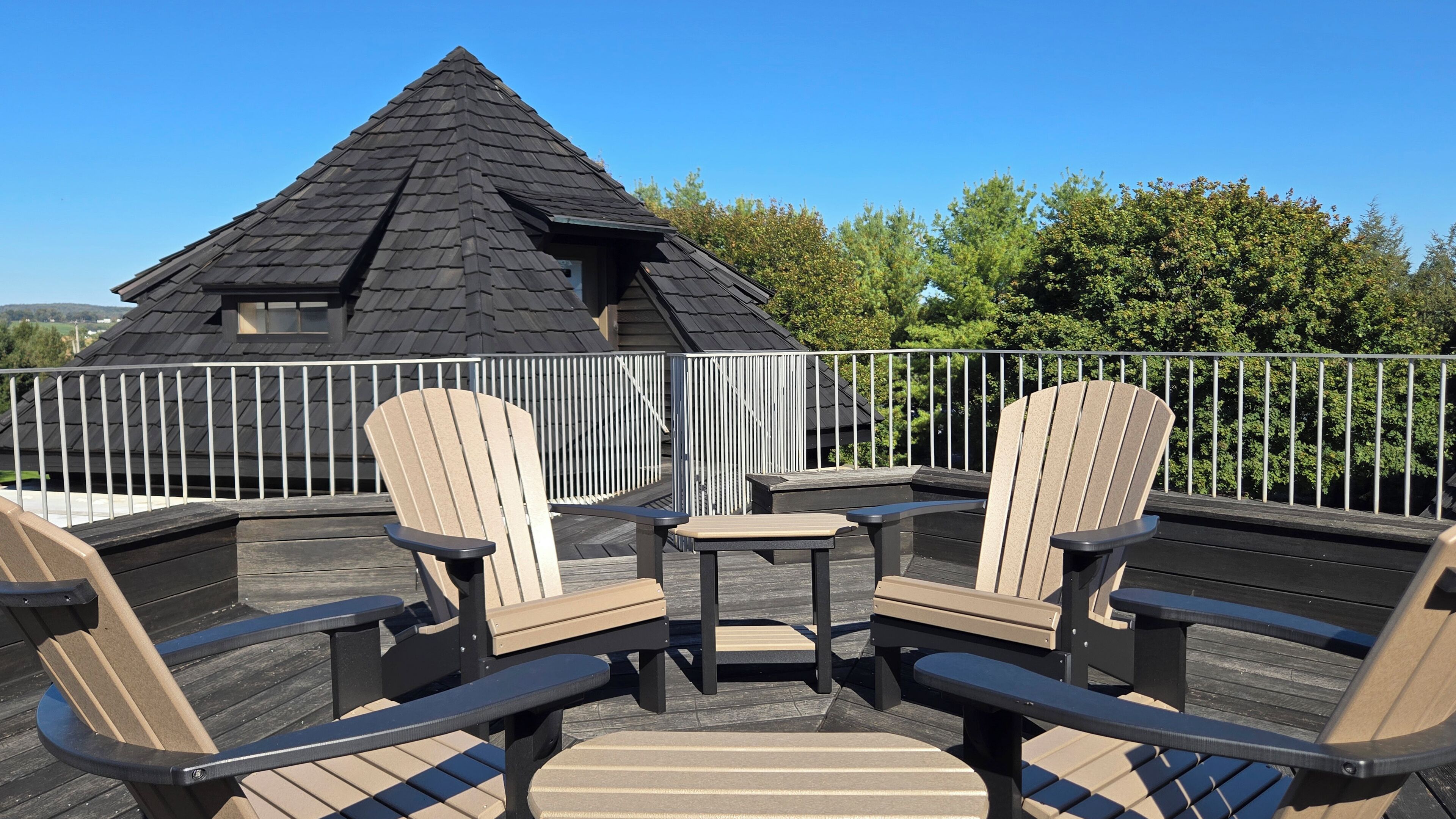 A deck with Adirondack chairs and a small table, surrounded by a white railing, overlooking a house with a dark roof.