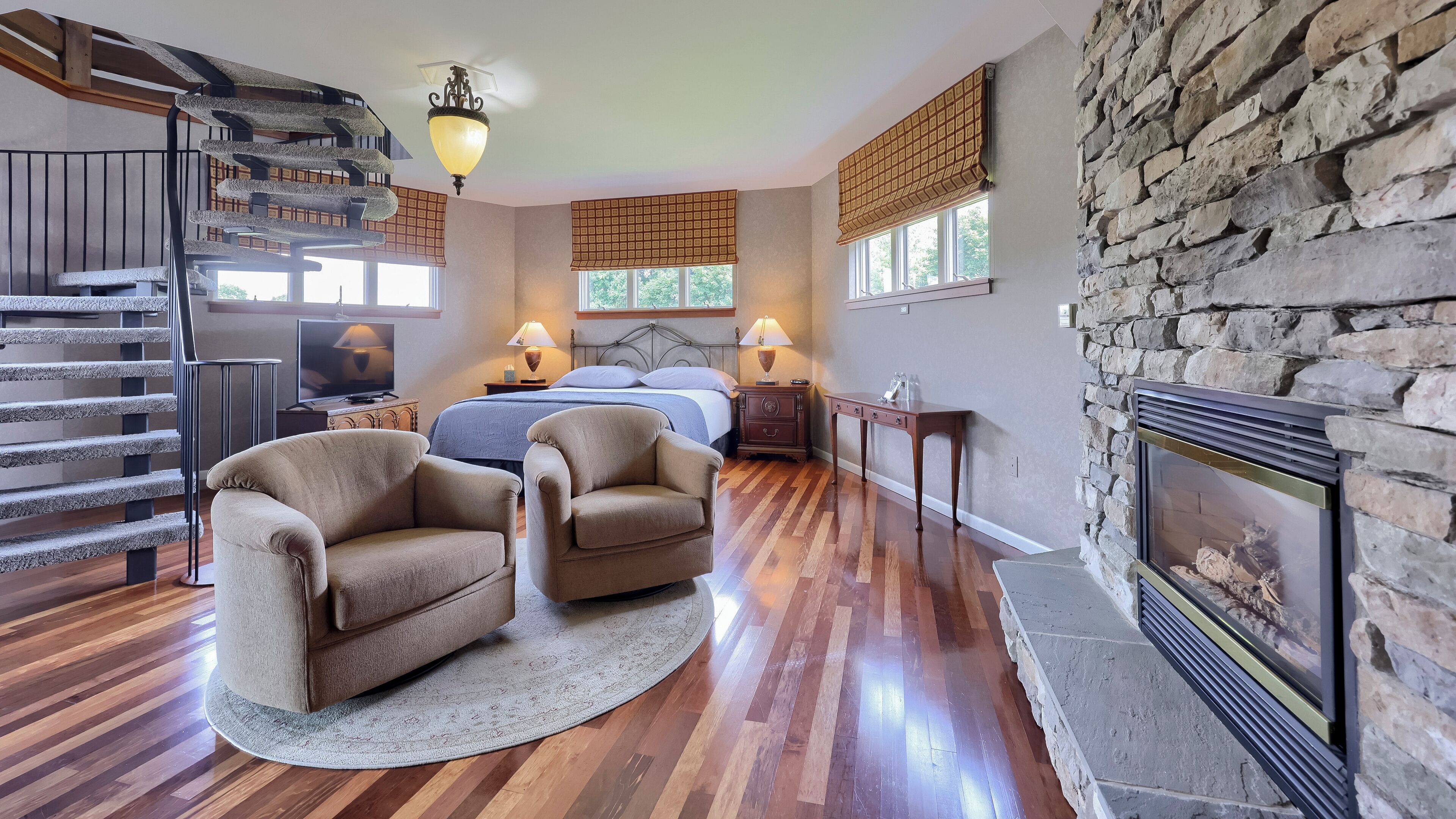 A bedroom with a stone fireplace, a bed with a headboard, a TV, and a staircase with a unique design.