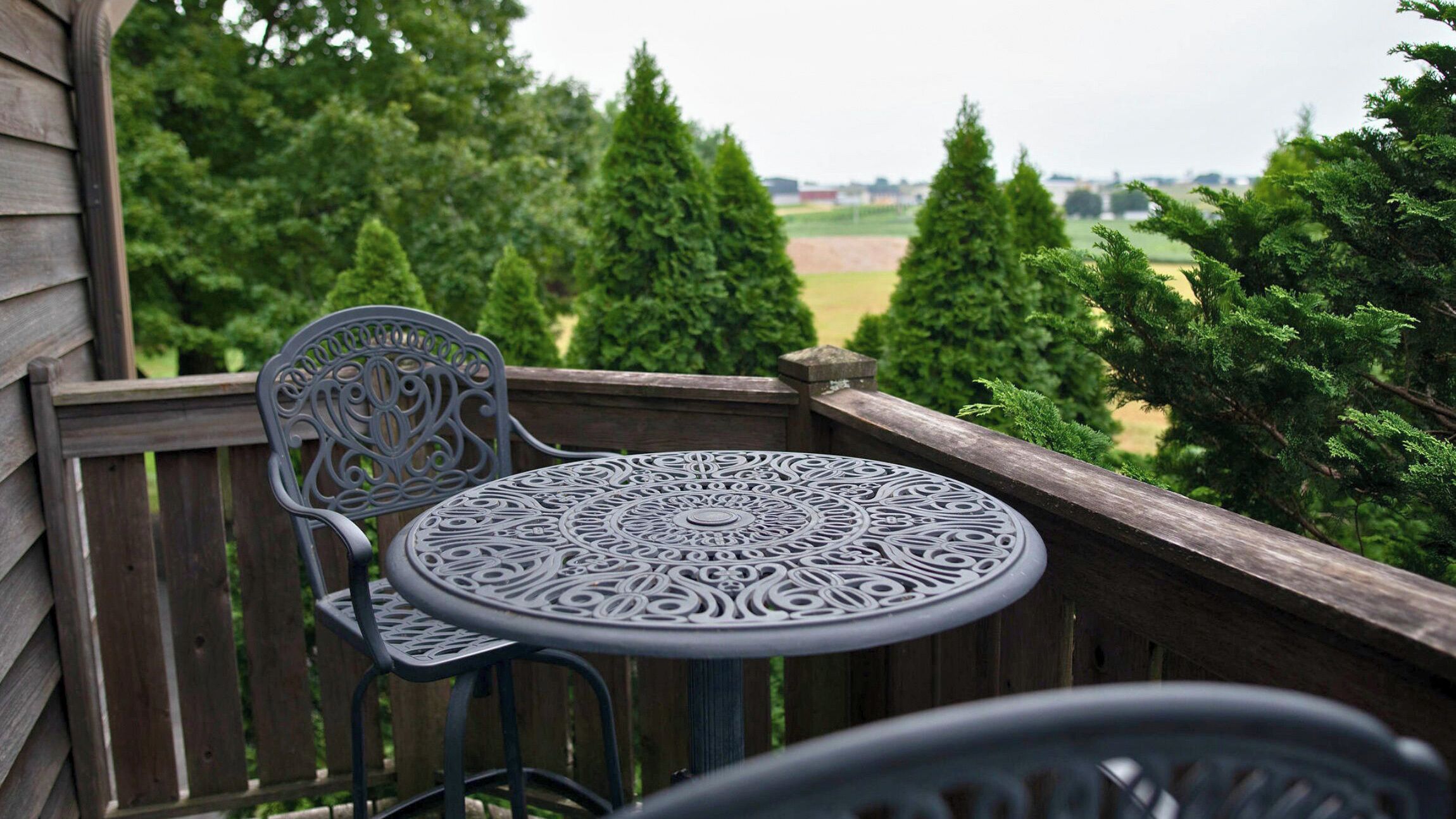 A wooden deck with a metal table and chair, overlooking a green landscape with trees.