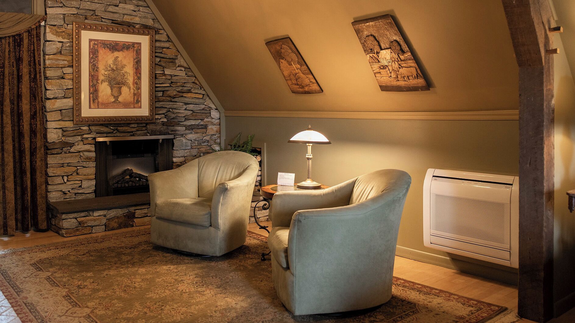 A cozy living room with a stone fireplace, two armchairs, a table with a lamp, and a radiator.