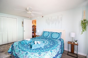 Un dormitorio con cama, ventilador de techo, mesita de noche con lámpara y colgador de macramé para planta en la pared.