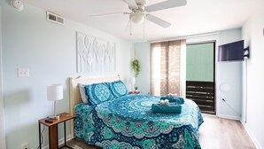 A bedroom with a bed, a ceiling fan, a wall-mounted TV, a window with blinds, and a plant. - #5 Steps away from beach! 1 mile from CB Boardwalk (Carolina Beach)