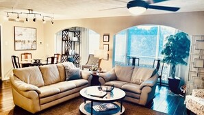 Ein Wohnzimmer mit einem Ecksofa, einem Couchtisch und einem Deckenventilator. Ein großes Fenster sorgt für natürliches Licht und einen Blick ins Freie.