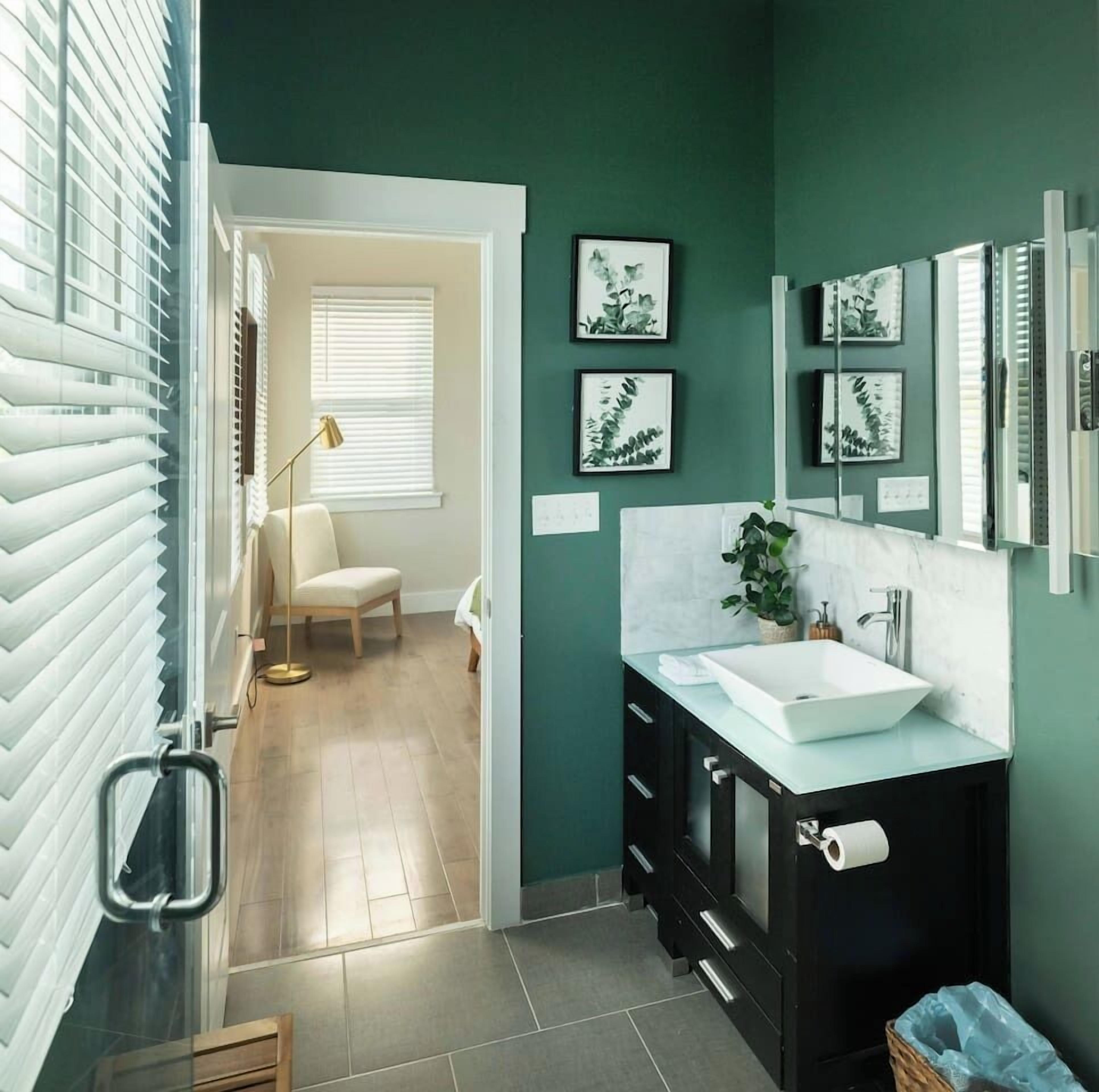 Un baño moderno con un lavamanos blanco, muebles oscuros y paredes verdes. Hay impresiones botánicas enmarcadas en la pared y una planta en maceta sobre el mostrador.