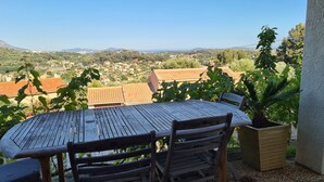 En udendørs terrasse med et træbord og stole, omgivet af krukker med planter og med udsigt over et boligområde med røde tegltage.
