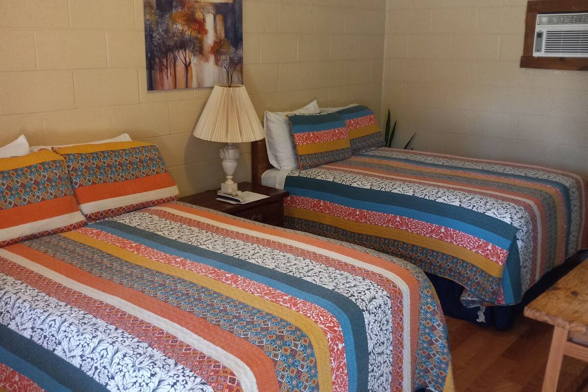 A room with two beds, a ceiling fan, a painting on the wall, and a wooden table.