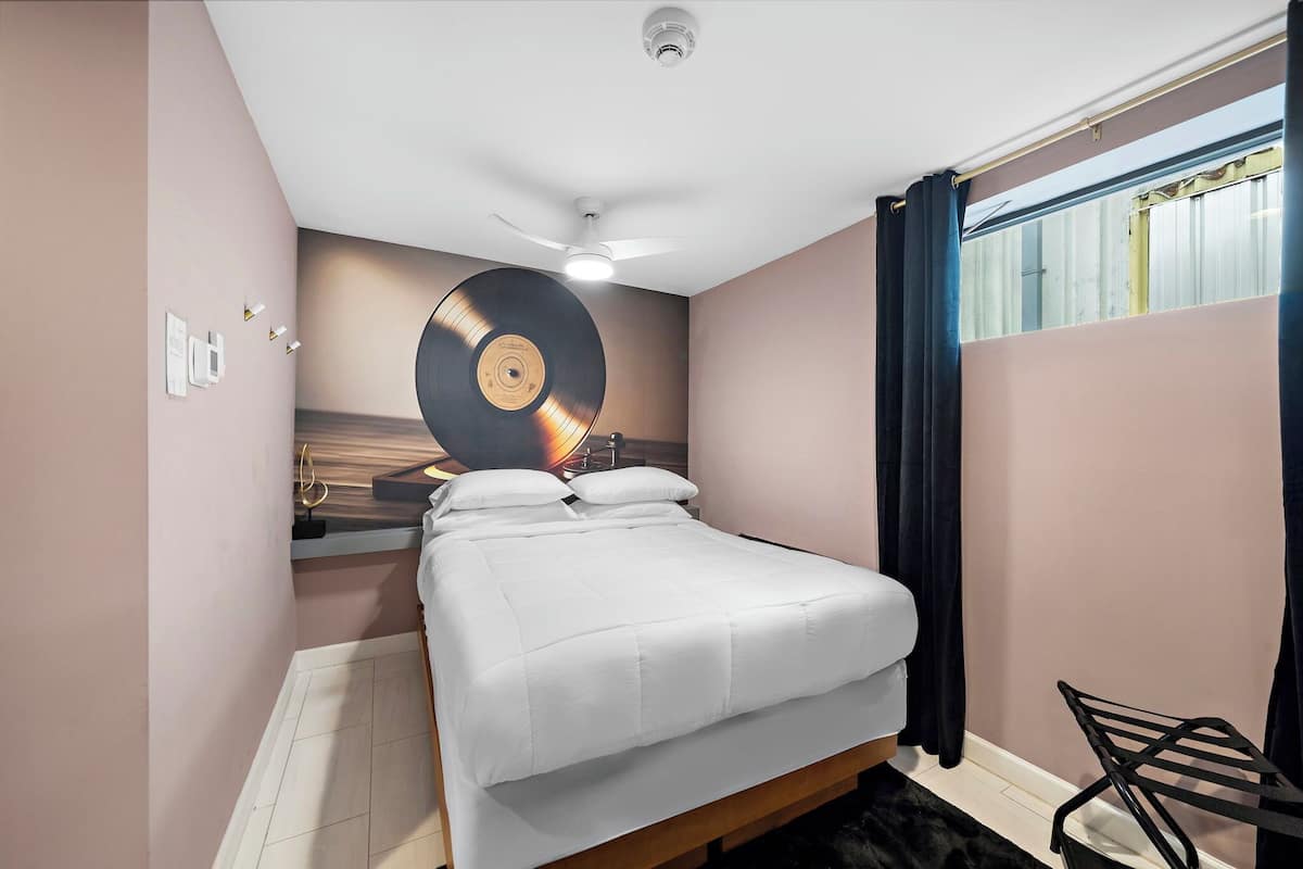 Een slaapkamer met een bed, een plafondventilator, een op de muur gemonteerde platenspeler en een raam met gordijnen.